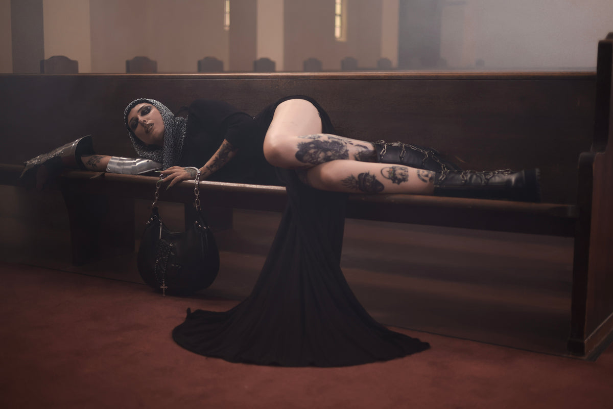 A person reclining on a wooden church pew in a dimly lit interior, wearing a long black dress, black knee-high boots, and a gray head covering. A black handbag rests beside them on the pew.